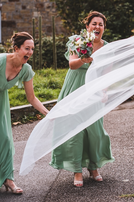 A documentary wedding photograph in Cornwall showing a bride's veil blowing in the wind while bridesmaids laugh.