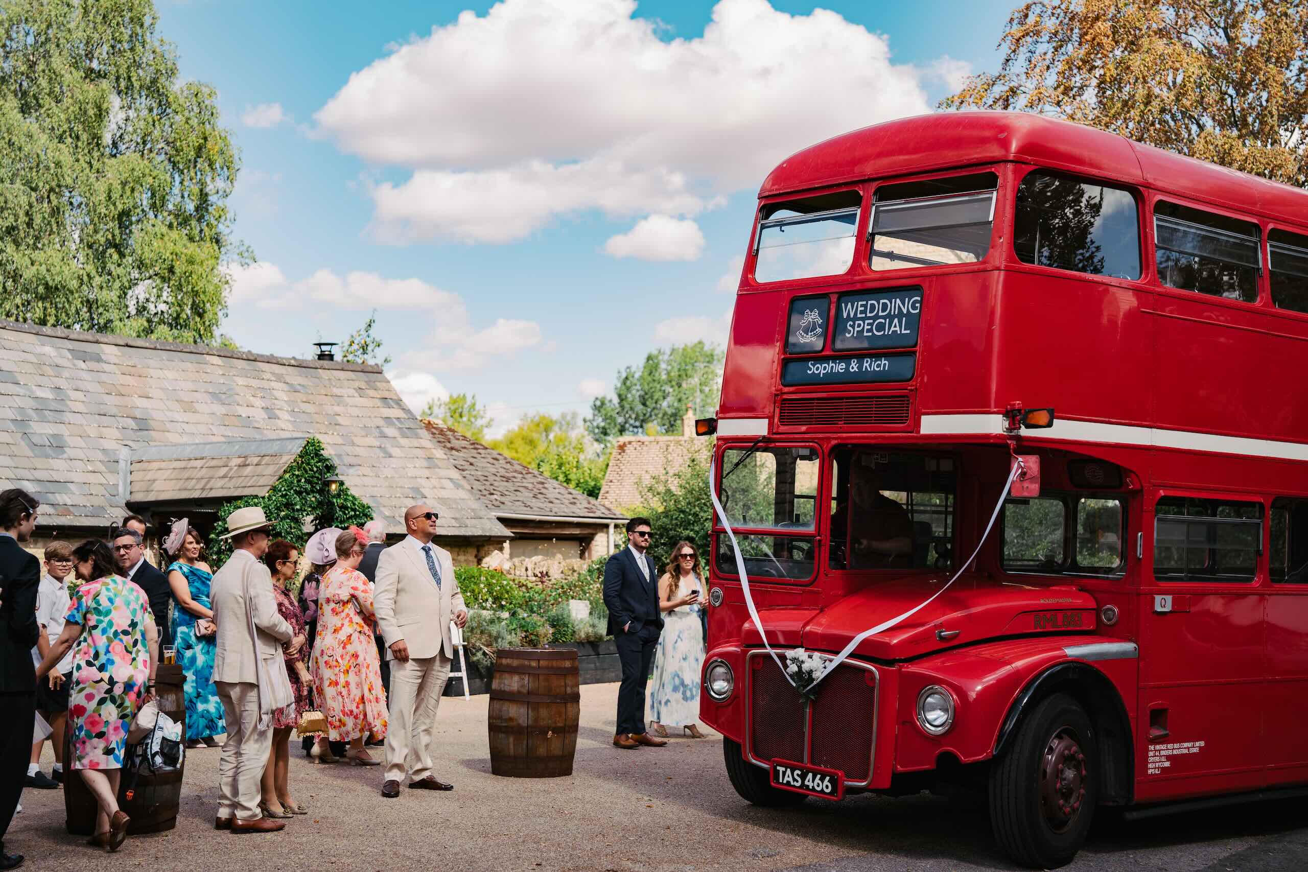 Wedding guests beside a vintage red bus at a Cotswolds countryside venue