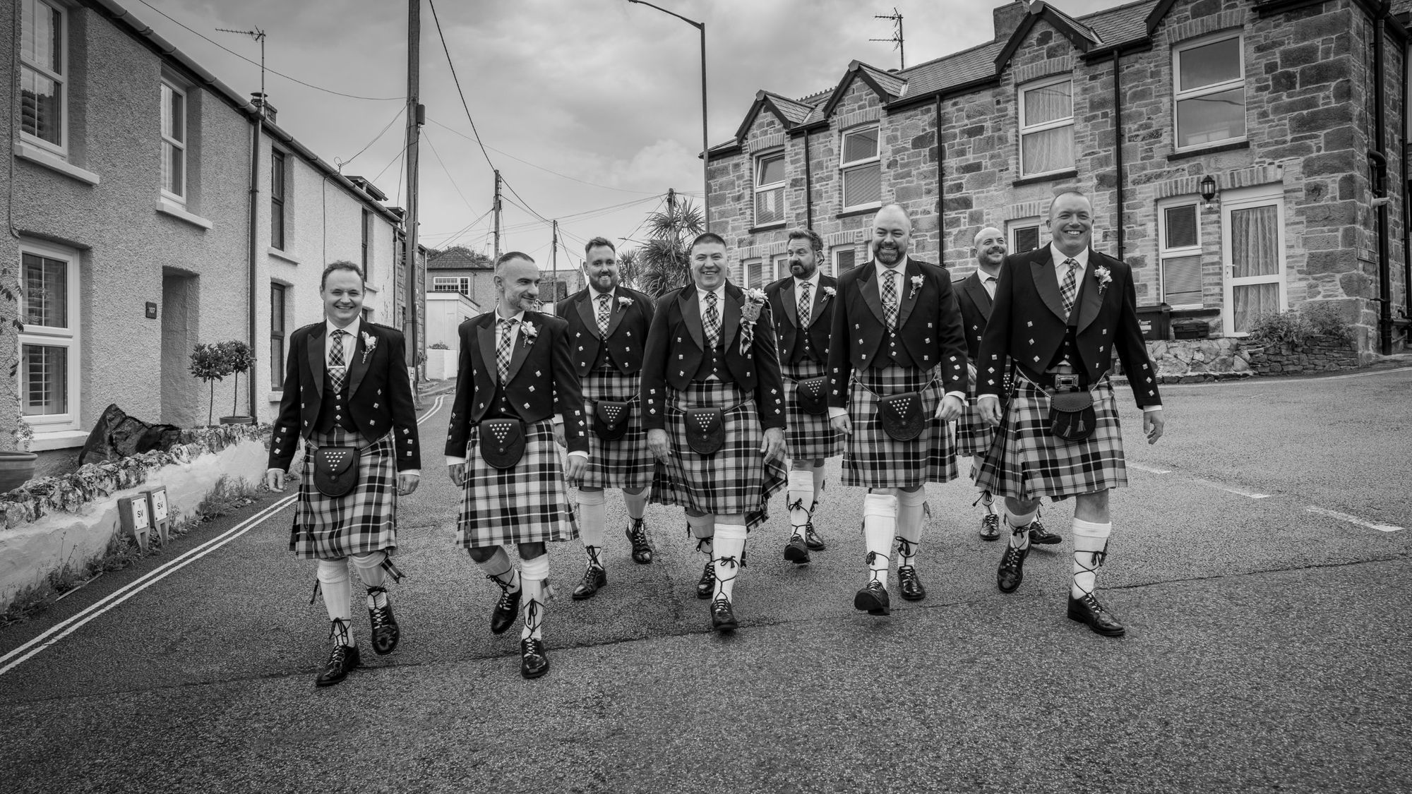Groom and groomsmen in kilts walking to St Columb Minor church, black and white