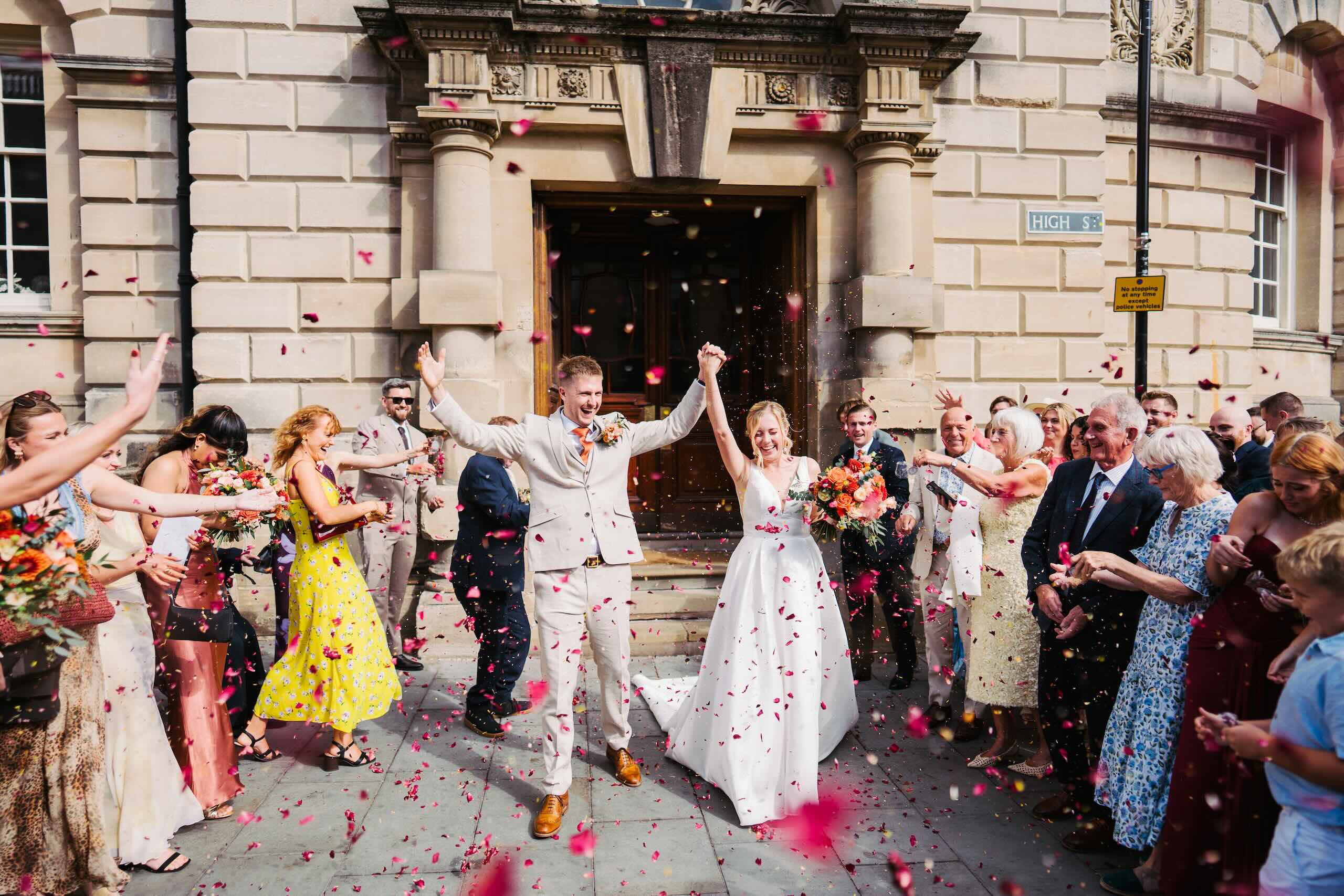 Bride and groom celebrating with confetti outside their ceremony venue, surrounded by cheering guests